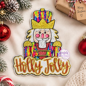Puede incluir: Un parche de lentejuelas con forma de cascanueces que presenta una corona dorada, una chaqueta arcoíris multicolor y una barba blanca. Debajo del cascanueces, el texto Holly Jolly está escrito en cursiva roja con borde dorado sobre fondo blanco.