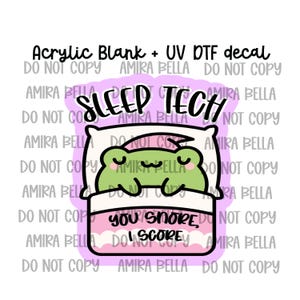 Peut inclure: Un support acrylique et un décalcomanie UV DTF représentant une grenouille verte dessinée dormant dans un lit avec un oreiller blanc et une couverture rose. Le texte indique SLEEP TECH en haut et YOU SNORE, I SCORE sur la couverture.