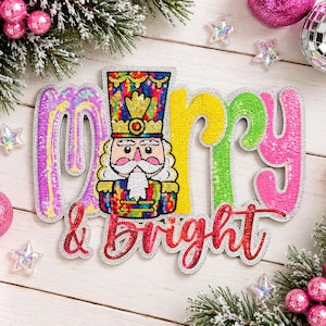 Könnte beinhalten: Eine Pailletten-Wanddekoration mit dem Schriftzug Merry & Bright, bei dem der Buchstabe M durch eine bunte Nussknacker-Figur ersetzt wird. Die Buchstaben sind mit leuchtenden violetten, gelben, grünen und rosa Pailletten besetzt.