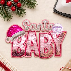Puede incluir: Un parche de lentejuelas termoadhesivo con el texto Santa Baby en rosa y plata. La palabra Santa está en letra cursiva plateada, mientras que Baby está en grandes letras de bloque rosas. Un gorro de Papá Noel rosa brillante está unido a la letra B.