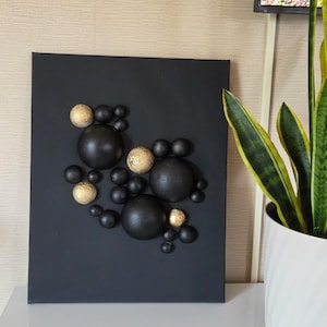 May include: A rectangular black canvas wall art piece featuring a cluster of three-dimensional spheres in varying sizes. The spheres are finished in matte black and textured metallic gold, arranged in an asymmetrical pattern across the surface.