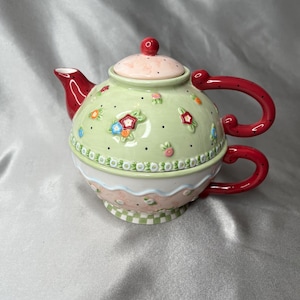 Puede incluir: Un juego de té individual de cerámica que incluye una tetera verde claro con asa y boquilla rojas de lunares, decorada con pequeñas flores en relieve y puntos negros. La base es una taza a juego con asa roja y un patrón de cuadros en la parte inferior.