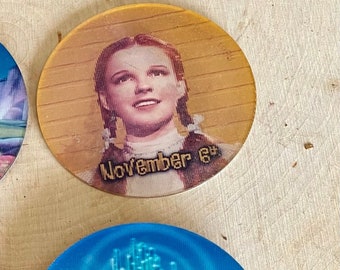 May include: Two round, colorful, Wizard of Oz themed coasters. One coaster features a picture of Dorothy from the movie with the text "November 6th". The other coaster features a picture of the yellow brick road leading to the Emerald City.