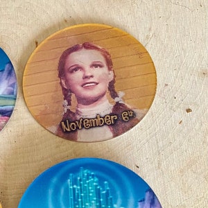 May include: Two round, colorful, Wizard of Oz themed coasters. One coaster features a picture of Dorothy from the movie with the text "November 6th". The other coaster features a picture of the yellow brick road leading to the Emerald City.