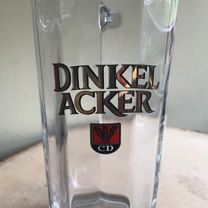 May include: A clear glass beer stein with a gold logo that says "Dinkel Acker" and a crest with the letters "CD" and three wheat stalks.
