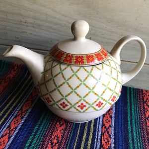 May include: White teapot with a red, green, and yellow geometric pattern. The teapot has a round body and a lid with a knob on top.