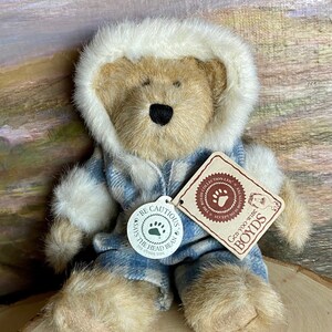 May include: A plush brown bear wearing a blue and white plaid jacket with a white fur-lined hood. The bear is sitting on a wooden surface. The bear has a tag that says "Gen-yoo-wine BOYDS" and "Be Cautious Says The Head Bean See Other Side".