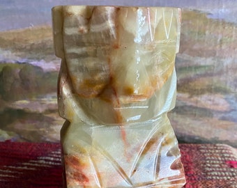 May include: A carved green and brown onyx statue of a person with a geometric design. The statue has a square base and stands on a striped rug.