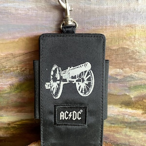 May include: Black fabric case with a silver-colored clip and a white cannon graphic. The text "AC/DC" is printed on a small patch at the bottom. The case has a loop on the back.