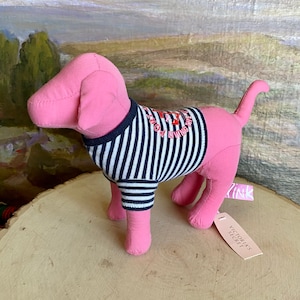 May include: Pink plush dog toy wearing a blue and white striped shirt with the text "Living the Dream" in red. The dog has a pink tag that says "PINK" and a white tag that says "Victoria's Secret".