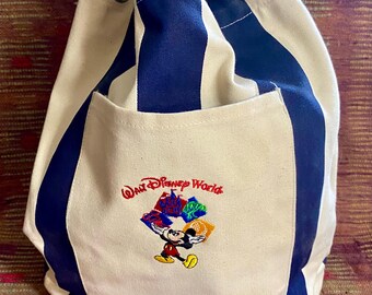 May include: Canvas drawstring bag with blue and white stripes. Features an embroidered image of Mickey Mouse carrying colorful gift bags, with the text "Walt Disney World". The bag has a front pocket and a drawstring closure.
