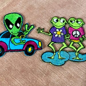 May include: Embroidered patches featuring a green alien in a blue car and two green frogs on a cloud. The alien is waving. The frogs wear shirts with a flower and peace sign.