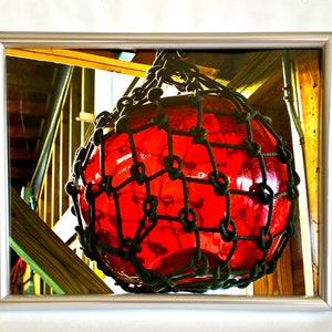 May include: A large, red glass buoy encased in a black rope net. The buoy is hanging from the ceiling of a wooden structure.