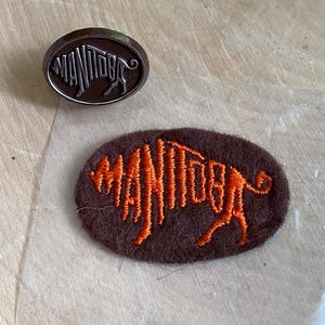 May include: An oval metal pin with a dark brown finish and the word Manitoba in raised silver lettering, paired with an oval brown felt patch featuring the word Manitoba embroidered in bright orange thread with a stylized animal tail design.