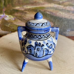 May include: A small, blue and white ceramic jar with a lid. The jar has a rounded body with a painted village scene and is supported by three small legs. The lid is topped with a small knob. The jar has two handles and is decorated with blue and black accents.