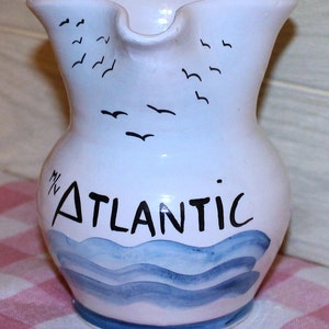 May include: White ceramic pitcher with a blue and white wave design and the text "M' ATLANTIC" in black. The pitcher has a handle and a spout.