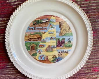 May include: A white ceramic plate with a gold trim and a detailed map of New Hampshire. The plate features a rope-like border and a colorful illustration of the state's landmarks, including a train, mountains, and various attractions.