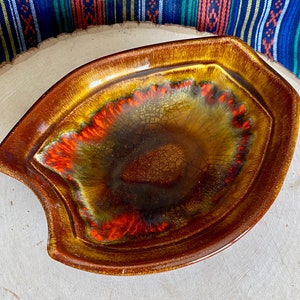 May include: A brown ceramic dish with a unique, abstract design featuring orange, yellow, and green swirls. The dish has a slightly wavy edge and a deep, rounded interior.