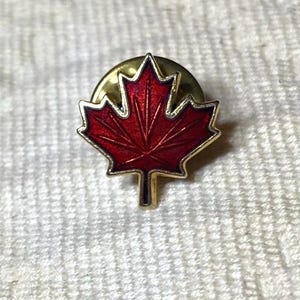 May include: A small lapel pin shaped like a maple leaf with a deep red enamel fill and a gold-toned metal border. The pin features a classic five-pointed leaf design with a central stem and a circular metal backing mechanism visible behind the leaf.