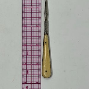 May include: A silver-toned metal button hook with a cream-colored handle, positioned next to a Westcott ruler showing a length of approximately 5 inches. The tool features a curved hook at the top and a ribbed metal section above the handle.