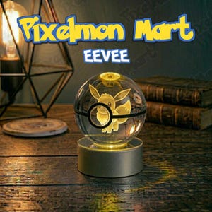 May include: A clear glass sphere featuring a laser-etched Eevee character inside, designed to resemble a Poké Ball with a black center band. The sphere rests on a cylindrical silver base and is illuminated from below, displaying the text Pixelmon Mart EEVEE.