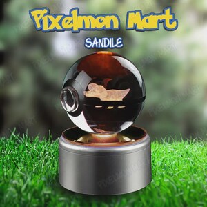 Puede incluir: Una esfera de cristal con una figura grabada de Sandile en su interior, sobre una base cilíndrica plateada. El texto Pixelmon Mart y Sandile aparece sobre la esfera. El diseño imita una bola con una banda central y un botón circular.