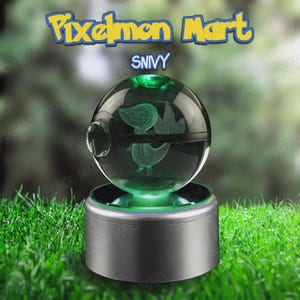Puede incluir: Una esfera de cristal transparente con forma de Poké Ball que contiene un personaje Snivy grabado con láser. La esfera descansa sobre una base cilíndrica plateada que emite luz verde. El texto Pixelmon Mart y Snivy aparece sobre el objeto.