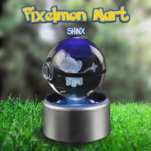 Puede incluir: Una esfera de cristal transparente con un personaje Shinx grabado con láser en su interior, similar a una Poké Ball con una banda negra. La esfera descansa sobre una base cilíndrica plateada que emite luz azul. El texto dice Pixelmon Mart y SHINX.
