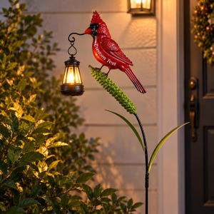 May include: A red cardinal bird garden stake featuring a black lantern hanging from its beak. The bird has the words Custom Text printed on its side and is perched on a green textured stem. The lantern emits a warm yellow light.