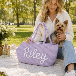 May include: A lavender-colored rope tote bag featuring the name Riley in white script lettering. The bag has two matching lavender rope handles and a structured, elongated shape, presented as a functional accessory for carrying personal items.