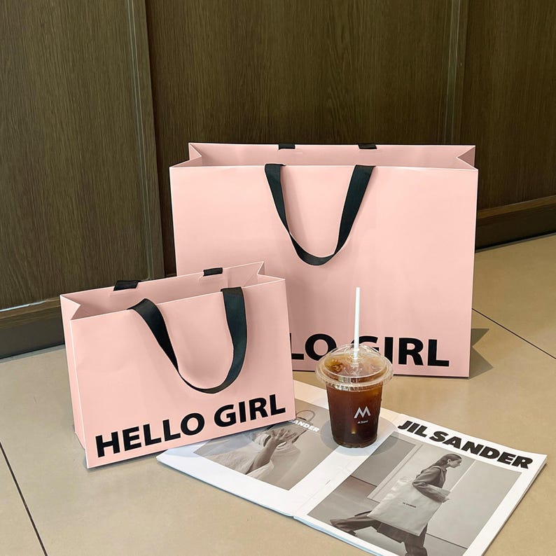 Puede incluir: Dos bolsas de papel de color rosa claro con asas de cinta negra y el texto HELLO GIRL impreso en negro. Un vaso de pl&aacute;stico transparente con caf&eacute; helado y una pajita blanca se sit&uacute;a frente a las bolsas junto a una revista Jil Sander abierta.