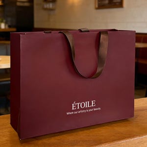 Puede incluir: Una bolsa de compras rectangular de papel color burdeos con asas de cinta marr&oacute;n oscuro. El frente presenta el texto blanco &Eacute;TOILE centrado sobre el texto m&aacute;s peque&ntilde;o Where our artistry is your beauty. Acabado mate y liso.