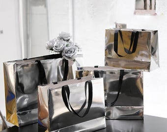 Puede incluir: Un conjunto de cuatro bolsas de regalo rectangulares de metal plateado con asas de cinta negra. Las bolsas tienen un acabado altamente reflectante, similar a un espejo. Un ramo de rosas artificiales plateadas se inserta en la bolsa.