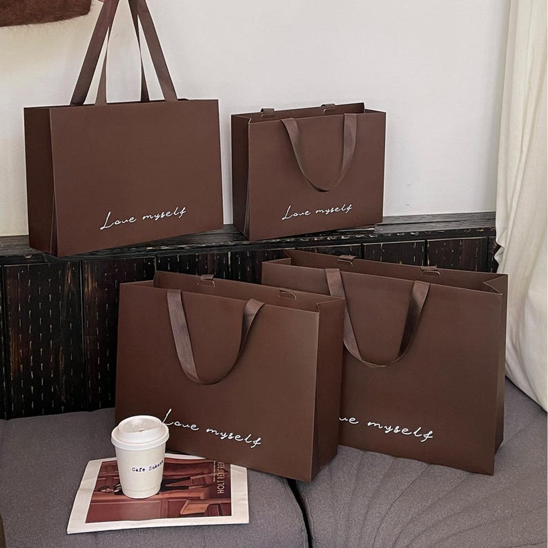 Puede incluir: Cuatro bolsas de compra rectangulares de papel marr&oacute;n oscuro con asas de cinta. Cada bolsa presenta el texto cursivo blanco "Love myself" impreso en el frente. Las bolsas est&aacute;n dispuestas en dos filas, con un vaso de caf&eacute; desechable blanco.