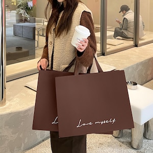 Puede incluir: Dos bolsas de compra rectangulares de papel marr&oacute;n oscuro con asas planas. La parte frontal de cada bolsa presenta el texto cursivo blanco 'Love myself' impreso cerca de la parte inferior. Las bolsas se sostienen juntas, mostrando un dise&ntilde;o limpio.