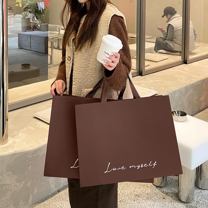 Puede incluir: Dos bolsas de compra rectangulares de papel marr&oacute;n oscuro con asas planas. La parte frontal de cada bolsa presenta el texto cursivo blanco 'Love myself' impreso cerca de la parte inferior. Las bolsas se sostienen juntas, mostrando un dise&ntilde;o limpio.