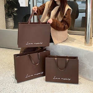 Puede incluir: Tres bolsas de compra rectangulares de papel marr&oacute;n oscuro con asas de cinta a juego. Cada bolsa presenta el texto cursivo blanco "Love myself" impreso en la parte frontal. Las bolsas est&aacute;n dispuestas en grupo, una sostenida y dos abajo.