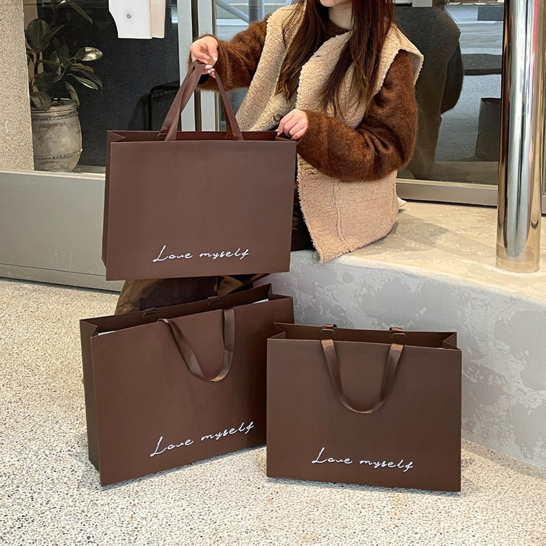 Puede incluir: Tres bolsas de compra rectangulares de papel marr&oacute;n oscuro con asas de cinta a juego. Cada bolsa presenta el texto cursivo blanco "Love myself" impreso en la parte frontal. Las bolsas est&aacute;n dispuestas en grupo, una sostenida y dos abajo.