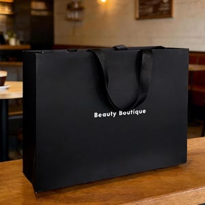 Puede incluir: Una bolsa de compras rectangular de color negro con asas de cinta negra. La parte frontal de la bolsa presenta el texto Beauty Boutique impreso en letras blancas sin remates. La bolsa tiene un acabado mate liso y una forma rectangular estructurada.