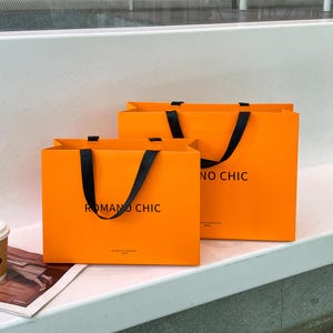 Puede incluir: Dos bolsas de compra rectangulares de color naranja con asas de cinta negra. En la parte frontal de cada bolsa aparece el texto negro ROMANO CHIC y un texto m&aacute;s peque&ntilde;o debajo que dice MAISON DE DOUCEUR PARIS. Las bolsas est&aacute;n una detr&aacute;s de otra.