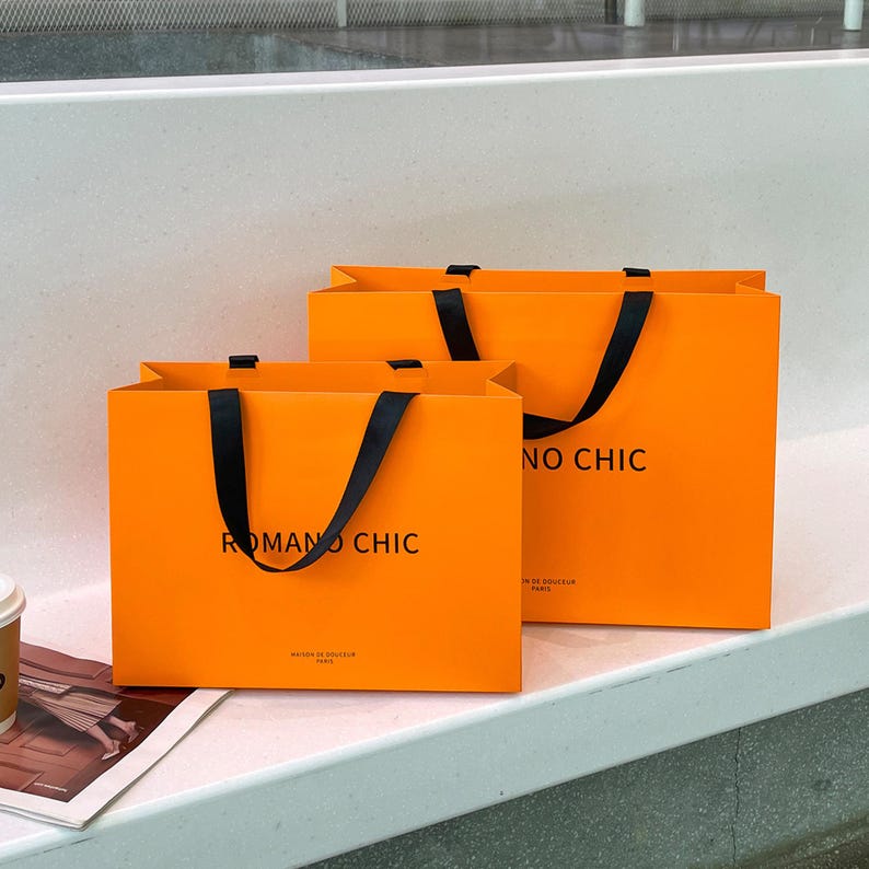 Puede incluir: Dos bolsas de compra rectangulares de color naranja con asas de cinta negra. En la parte frontal de cada bolsa aparece el texto negro ROMANO CHIC y un texto m&aacute;s peque&ntilde;o debajo que dice MAISON DE DOUCEUR PARIS. Las bolsas est&aacute;n una detr&aacute;s de otra.