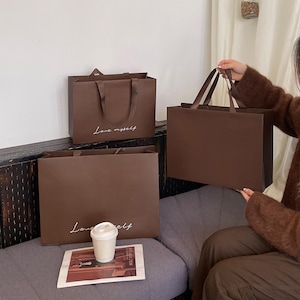 Puede incluir: Tres bolsas de compra rectangulares de papel marr&oacute;n oscuro con asas de cinta. Dos de las bolsas presentan el texto cursivo blanco "Love myself" impreso en la parte frontal. Las bolsas est&aacute;n fabricadas con un material mate y tienen una forma estructurada.