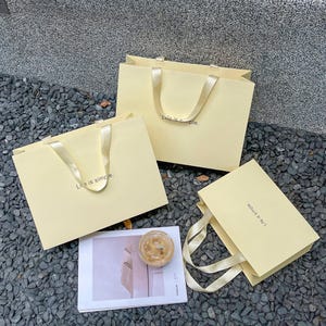 Puede incluir: Tres bolsas de regalo de papel amarillo p&aacute;lido con asas de cinta de sat&eacute;n a juego. Cada bolsa presenta el texto "Life is simple." impreso en una fuente peque&ntilde;a y oscura en el frente. Las bolsas est&aacute;n dispuestas junto a una tarjeta rectangular.