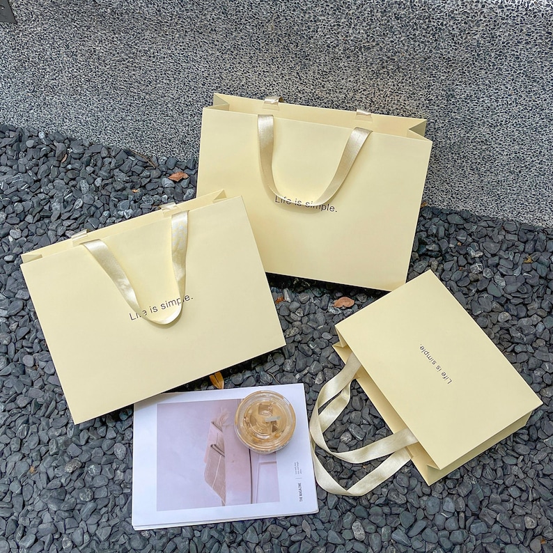 Puede incluir: Tres bolsas de regalo de papel amarillo p&aacute;lido con asas de cinta de sat&eacute;n a juego. Cada bolsa presenta el texto "Life is simple." impreso en una fuente peque&ntilde;a y oscura en el frente. Las bolsas est&aacute;n dispuestas junto a una tarjeta rectangular.