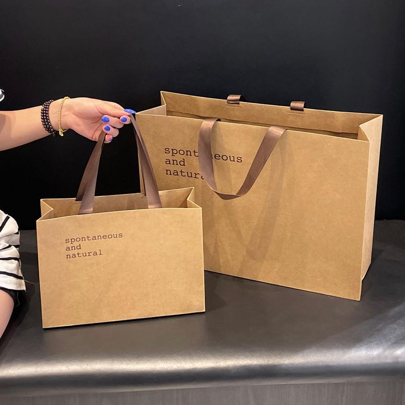 Puede incluir: Dos bolsas de compra rectangulares de papel marr&oacute;n con asas de cinta marr&oacute;n oscuro. Cada bolsa presenta el texto impreso en negro "spontaneous and natural" en la parte frontal. Las bolsas est&aacute;n hechas de papel liso con acabado mate.