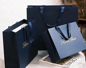 Puede incluir: Tres bolsas de papel de color azul marino oscuro con asas de cinta azul marino a juego. Cada bolsa presenta el texto Rara Gaax impreso en color dorado en la parte frontal. Las bolsas están dispuestas en grupo, con dos pequeñas frente a una grande.