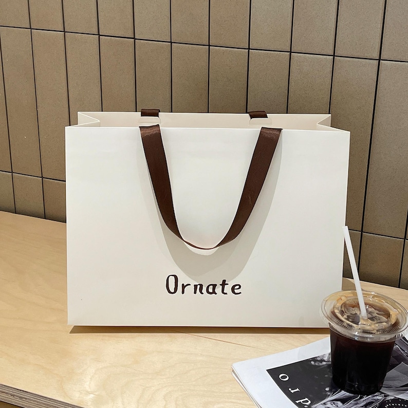 Puede incluir: Una bolsa de papel rectangular de color blanco roto con asas de cinta marr&oacute;n oscuro y la palabra Ornate impresa en una fuente sans-serif marr&oacute;n oscuro en el centro. La bolsa tiene un dise&ntilde;o limpio y estructurado con una base plana.
