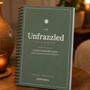 May include: A sage green spiral-bound planner with white text that reads: THE Unfrazzled PLANNER, an ADHD-friendly daily system made to work with your brain, not against it, UNDATED - USE ANY YEAR, and driftwoodery, featuring a small white circle.
