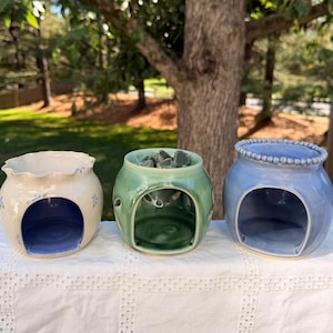 May include: Three ceramic wax warmers arranged in a row. The left warmer is cream with blue floral accents and a ruffled rim. The center warmer is sage green with dark green wax melts inside. The right warmer is light blue with a beaded rim.