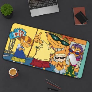 May include: A rectangular desk mat featuring a colorful cartoon illustration of four characters from the show Rocket Power. The design includes the text THE PIE on a blue sign, with characters wearing casual clothing like shorts, shirts, and sunglasses.